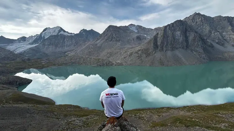 Da dietro, un capo gruppo WeRoad siede su una roccia che si affaccia su un lago alpino turchese che riflette montagne innevate.