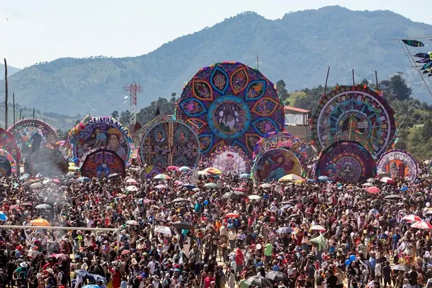 Una grande folla si raduna per un festival con aquiloni giganti, colorati e circolari, con montagne verdi sullo sfondo.