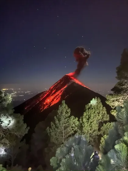 Una persona di un viaggio di gruppo WeRoad osserva un vulcano in eruzione di notte, con lava rossa che scorre e una colonna di fumo contro un cielo stellato.