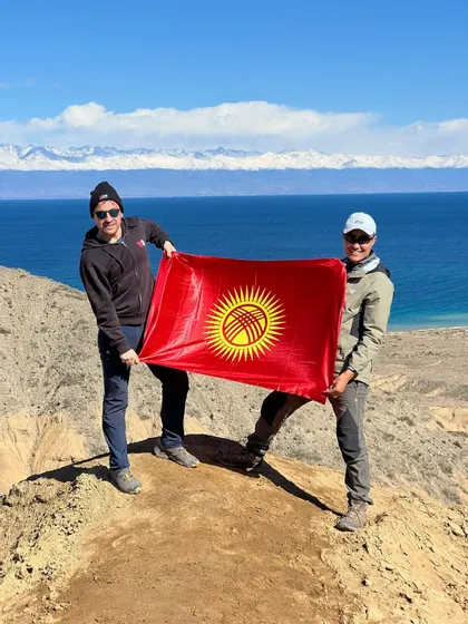 Due uomini di un viaggio di gruppo WeRoad tengono la bandiera del Kirghizistan su una collina che domina un lago e montagne innevate.
