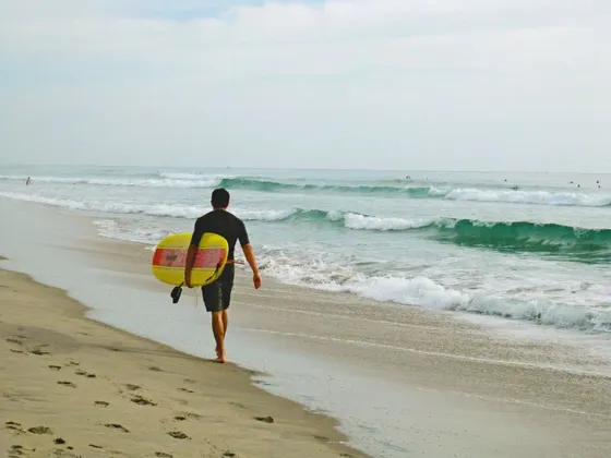 Un surfista in muta cammina lungo una spiaggia sabbiosa, trasportando una tavola da surf gialla verso il mare.