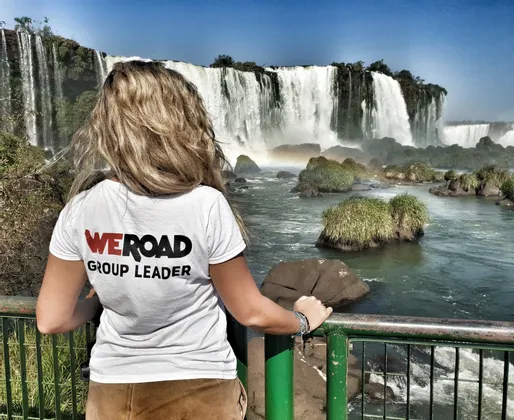 Eine blonde WeRoad Gruppenleiterin steht an einem Geländer und blickt auf einen großen, kaskadierenden Wasserfall unter klarem blauem Himmel.