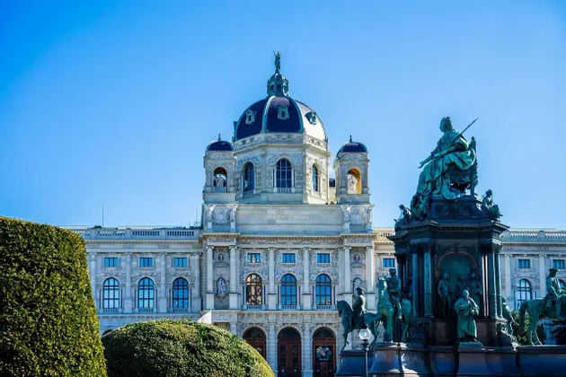 Un bâtiment historique orné d'un grand dôme se dresse derrière un monument imposant avec plusieurs statues sous un ciel bleu clair.