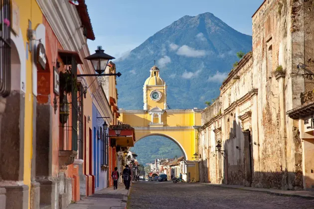 Un arco giallo con una torre dell'orologio si erge su una strada di ciottoli, con un grande vulcano che si staglia sullo sfondo.