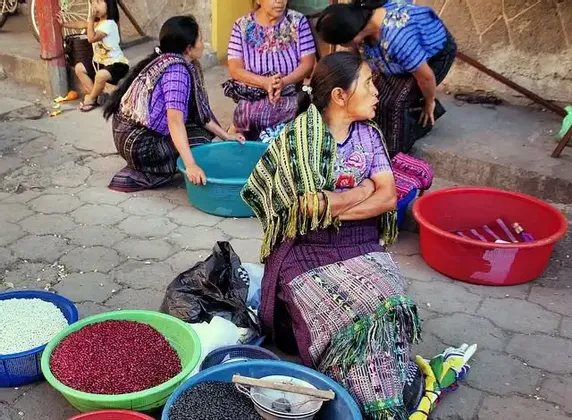 Un gruppo di donne in abiti tradizionali siede su una strada lastricata vendendo fagioli colorati da grandi bacinelle.