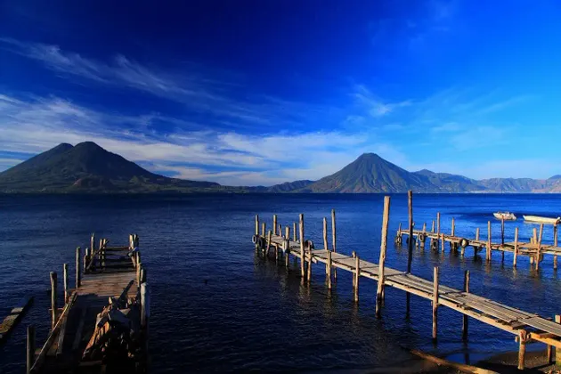 Pontili in legno si estendono in un lago blu calmo con grandi montagne sullo sfondo sotto un cielo parzialmente nuvoloso.