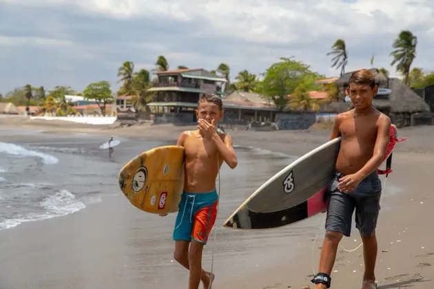 Deux jeunes garçons avec des planches de surf marchent le long du rivage sur une plage de sable avec des palmiers en arrière-plan.