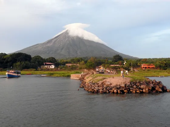 Un volcan avec un nuage lenticulaire sur son sommet surplombe un plan d'eau avec un bateau et une jetée rocheuse au premier plan.