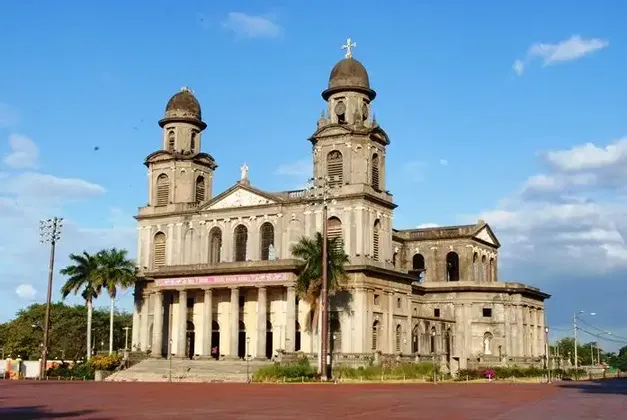 Une grande cathédrale de pierre avec deux clochers à dôme et une entrée à colonnes, vue depuis une place en briques rouges.
