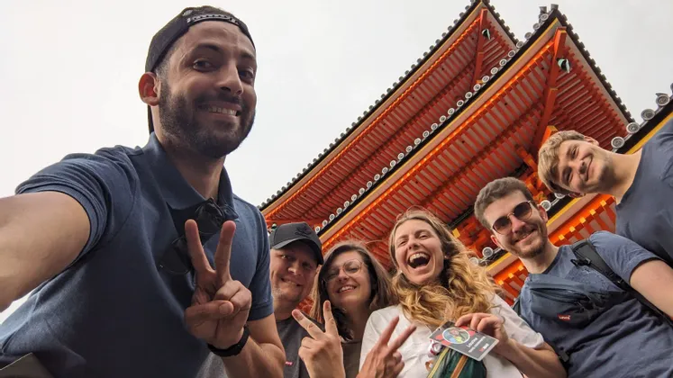 Un groupe WeRoad prend un selfie en contre-plongée, souriant et faisant des signes de paix devant un temple japonais rouge.