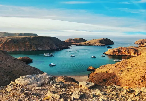 Una vista aerea di diverse barche ancorate in una baia turchese, circondata da aride colline rocciose sotto un cielo azzurro.