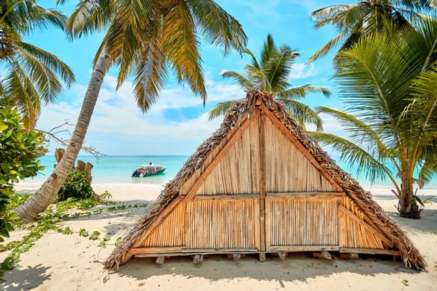 Eine dreieckige Holzhütte mit Strohdach steht an einem weißen Sandstrand unter Palmen, mit einem Boot auf dem türkisfarbenen Wasser.
