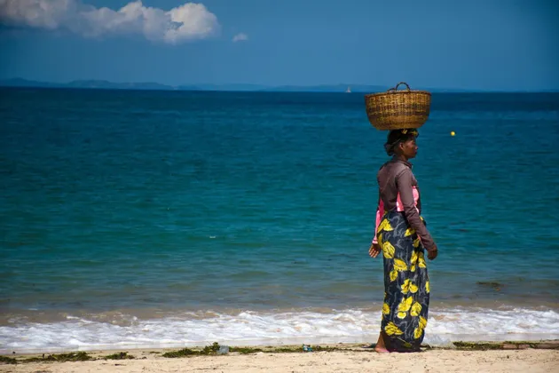 Eine Frau geht an einem Sandstrand am blauen Ozean entlang und balanciert einen großen Weidenkorb auf ihrem Kopf unter klarem Himmel.
