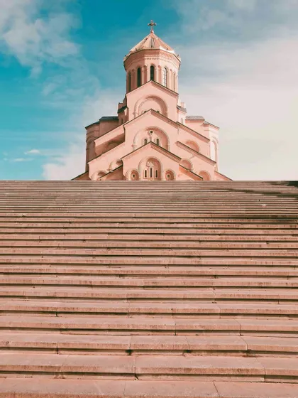 Una vista dal basso di un'ampia scalinata in pietra che porta a una grande chiesa di colore rosato con una cupola contro un cielo blu.
