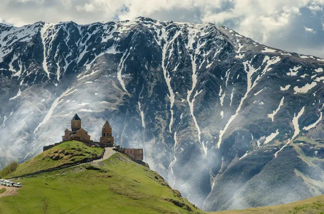 Un'antica chiesa in pietra con torri, su una verde collina con un'imponente montagna striata di neve sullo sfondo.