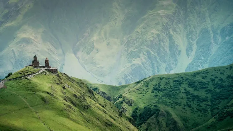 Un complesso monastico in pietra sorge su una rigogliosa collina verde, con uno sfondo di imponenti montagne nebbiose.