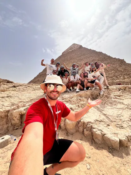 Ein Mann macht ein Selfie mit seiner WeRoad-Gruppe, die gemeinsam auf Felsen vor einer großen Pyramide unter strahlend blauem Himmel posiert.