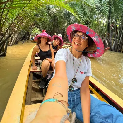 Eine WeRoad-Gruppe von Frauen mit pinkfarbenen Spitzhüten macht ein Selfie auf einem Boot in einem von Palmen gesäumten Kanal.
