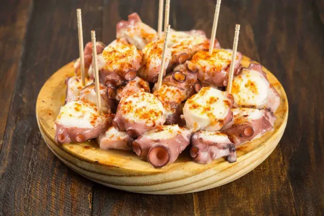 Trozos de pulpo cocido sazonados con pimentón, servidos con palillos en un plato de madera redondo.