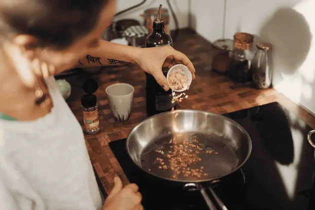 Vista por encima del hombro de una persona con tatuaje añadiendo ingredientes picados de un bol a una sartén caliente en una placa de cocción