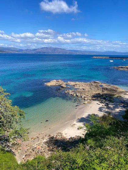 Una vista costera panorámica de una pequeña playa de arena anidada entre rocas, con agua turquesa y montañas en el horizonte bajo un cielo azul.