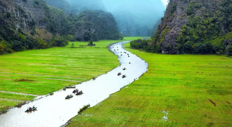 Un viaje en grupo de WeRoad en pequeñas embarcaciones navega por un río serpenteante a través de campos verdes vibrantes, enclavado entre grandes montañas rocosas.