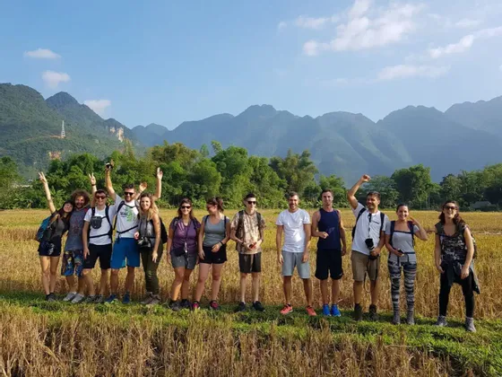 Eine WeRoad Gruppe posiert für ein Foto auf einem goldenen Feld vor grünen Bergen unter blauem Himmel.