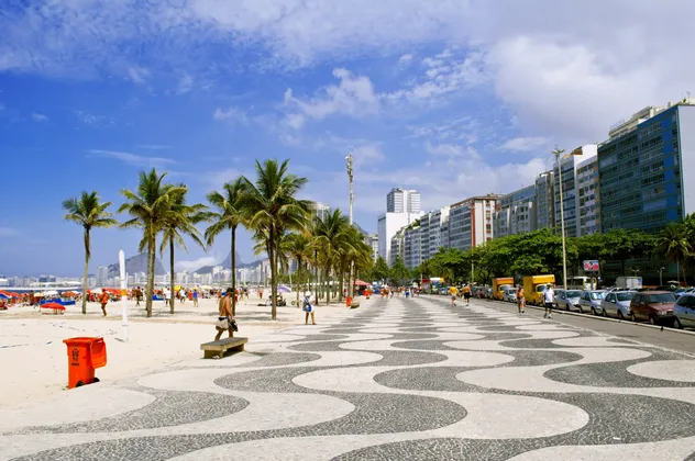 Une promenade en mosaïque à motifs de vagues longe une plage de sable bordée de palmiers, avec la ligne d'horizon de la ville en arrière-plan.