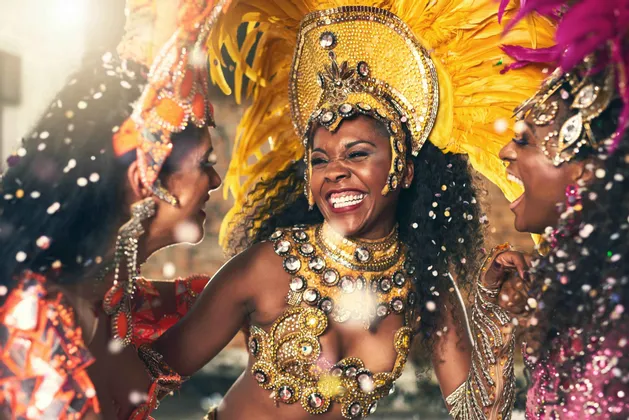Tres mujeres con elaborados trajes de carnaval emplumados ríen y bailan juntas entre confeti cayendo.