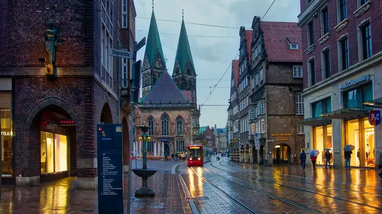 Un tranvía rojo viaja por vías mojadas a través de una calle de ciudad europea al anochecer, con una catedral de dos agujas al fondo.