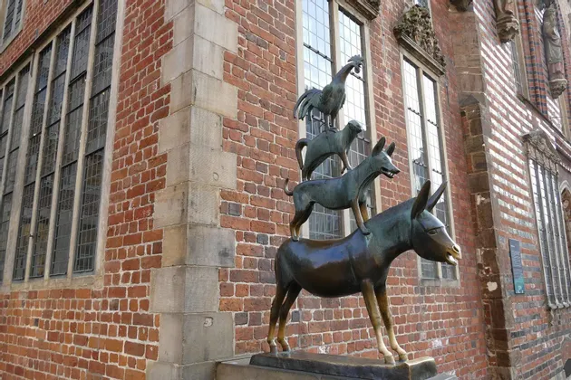 Una estatua de bronce de un burro, perro, gato y gallo apilados uno encima del otro junto a un edificio de ladrillo rojo.