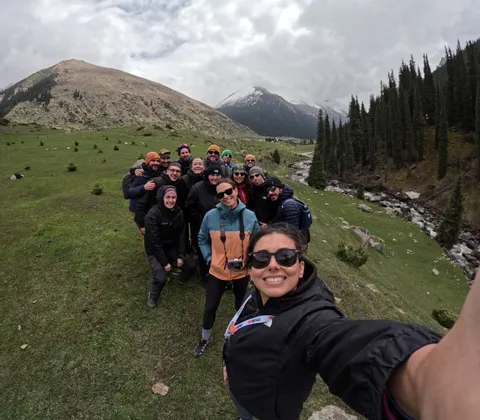 A WeRoad group trip poses for a selfie on a grassy mountainside, with a river and snow-capped peaks in the background.