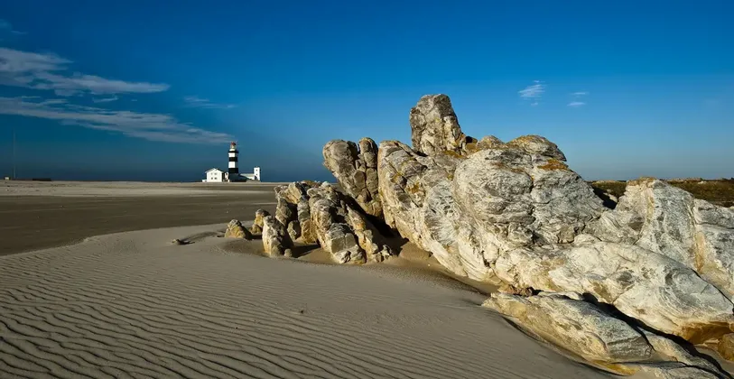 Una gran formación rocosa se encuentra en una playa de arena rizada, con un faro blanco y negro a lo lejos bajo un cielo azul.