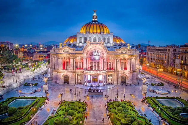 Edificio con cúpula ornamentada iluminado al atardecer, visto desde arriba, con jardines cuidados y una plaza pública en primer plano.