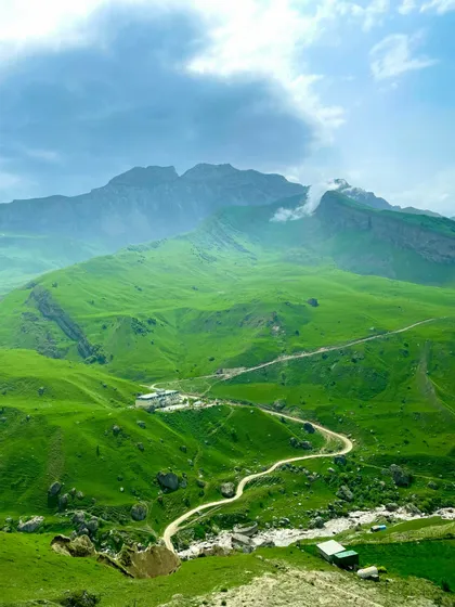 Una strada sterrata tortuosa attraversa una valle di colline verdeggianti con edifici sparsi, sotto una grande catena montuosa rocciosa.
