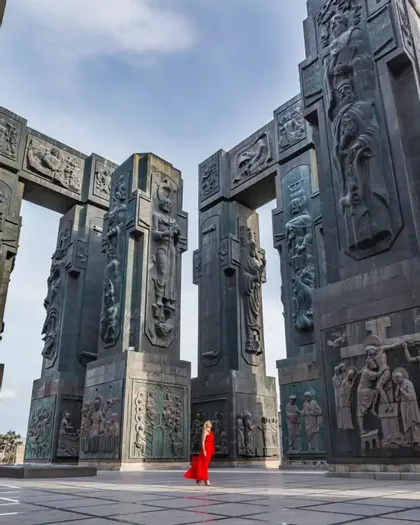 Una donna con un lungo abito rosso si erge davanti a colossali monumenti in pietra scolpita in un'ampia piazza aperta.