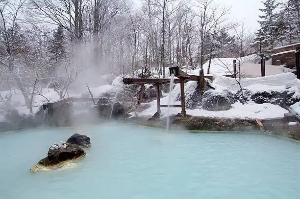 Un acquedotto di legno riversa acqua in una sorgente termale fumante e lattiginosa, di colore azzurro, in un paesaggio innevato e boscoso.