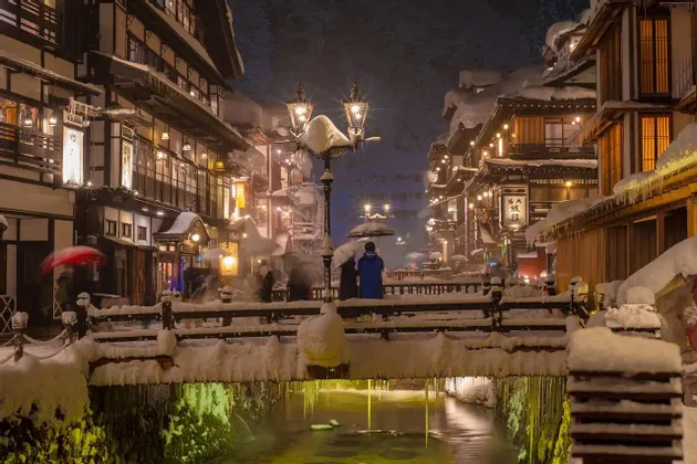Un villaggio innevato di notte, con edifici tradizionali, un ponte su un canale e lanterne illuminate.