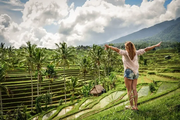 Una mujer con los brazos extendidos se alza sobre una colina, contemplando terrazas de arroz verdes y palmeras bajo un cielo nublado.