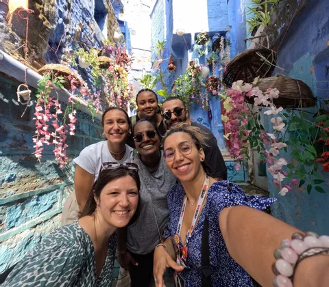 Sechs Personen einer WeRoad-Gruppenreise lächeln für ein Selfie in einer engen Gasse mit leuchtend blauen Wänden und hängenden Blumen.