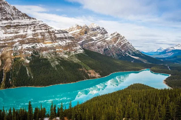 Ein leuchtend türkiser See spiegelt schneebedeckte Berge wider, umgeben von dichtem Nadelwald unter einem teils bewölkten Himmel.