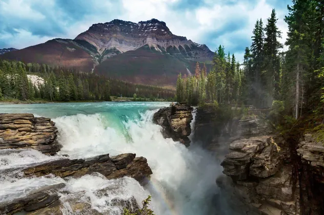 Ein türkisblauer Fluss stürzt über einen breiten, felsigen Wasserfall, im Hintergrund ein Kiefernwald und ein großer Berg.