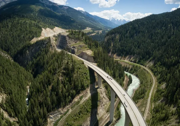 Luftaufnahme einer Autobahnbrücke mit Autos, die sich über ein bewaldetes Tal mit einem Fluss und Bergen schlängelt.