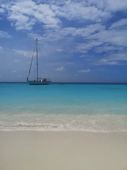 Una barca a vela ancorata in acque calme e turchesi, vista da una spiaggia di sabbia bianca sotto un cielo parzialmente nuvoloso.