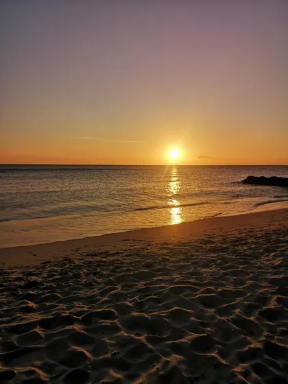 Il sole tramonta all'orizzonte sull'oceano, proiettando un riflesso dorato sull'acqua di fronte a una spiaggia sabbiosa.