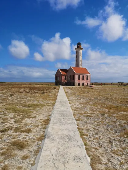 Un lungo sentiero di cemento conduce a un faro logoro e a una costruzione annessa in un paesaggio piatto e arido sotto un cielo azzurro.