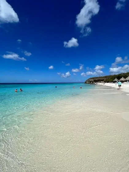 Persone che nuotano in acqua cristallina turchese su una spiaggia di sabbia bianca sotto un cielo blu intenso con nuvole bianche.