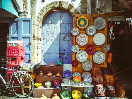 Una bancarella del mercato espone ceramiche colorate, tajine e borse in pelle davanti a una grande porta di legno blu rustica.