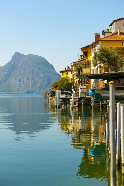 Edifici colorati sulla riva di un lago si riflettono nell'acqua calma, con una grande montagna che si erge sullo sfondo.