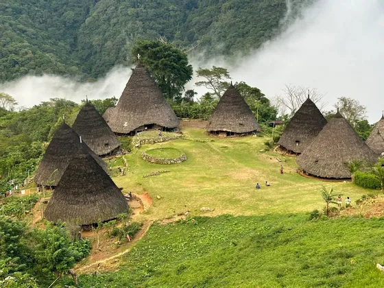 Un village traditionnel avec des huttes coniques aux toits de chaume sur une colline verdoyante, entouré de montagnes boisées et de nuages bas.